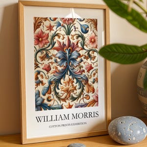 May include: A framed print with a detailed floral design in red, blue, and orange hues. The print is titled "WILLIAM MORRIS COTTON PRINTS EXHIBITION" in black lettering. The frame is made of light-toned wood, typical of a gallery style.