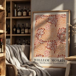 Op de afbeelding: Een ingelijste print met een herhalend patroon van gestileerde bladeren en bloemelementen in tinten rood, wit en perzik. De print is getiteld "WILLIAM MORRIS" en "the Smithsonian Institution". De print wordt getoond in een kamer met een houten boekenplank en een comfortabele fauteuil.