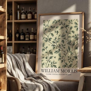 Op de afbeelding: Een ingelijste print met een klassiek textielpatroonontwerp van William Morris. Het kunstwerk toont een herhalend patroon van groene bladeren en wijnstokken op een lichtgele achtergrond. De print staat in een kamer met een houten boekenplank en een comfortabele stoel.