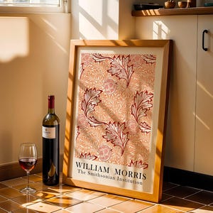 Op de afbeelding: Een ingelijste William Morris-print met een bloemmotief in rood, crème en beige. Een fles rode wijn en een wijnglas met rode wijn staan voor de print. De print is getiteld "William Morris The Smithsonian Institution."