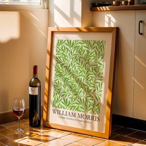 May include: A framed print with a green leaf pattern and the text "WILLIAM MORRIS CLASSIC TEXTILE PATTERN DESIGN" leans against a wall. A bottle of red wine and a wine glass with red wine are in front of the print.