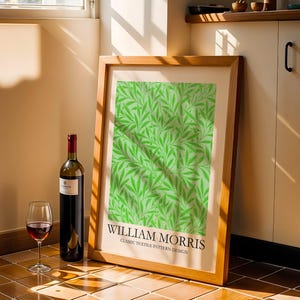 Op de afbeelding: Een ingelijste print met een groen bladerpatroon en de tekst "WILLIAM MORRIS CLASSIC TEXTILE PATTERN DESIGN". Een fles rode wijn en een wijnglas staan voor de print.