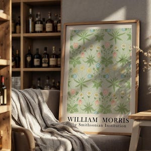 Op de afbeelding: Een ingelijste print met een bloemenpatroon in pastelkleuren, met de tekst "WILLIAM MORRIS the Smithsonian Institution." De print wordt getoond in een kamer met een houten plank vol flessen en een comfortabele fauteuil.