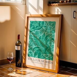 Op de afbeelding: Een ingelijste print van een klassiek textielpatroon van William Morris in groentinten, leunend tegen een muur. Een fles rode wijn en een wijnglas met rode wijn staan voor de print. De print heeft een houten lijst.
