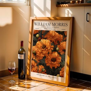 May include: A framed William Morris art print with orange floral design, a wine bottle, and a wine glass with red wine. The print leans against a wall, and the scene is lit by sunlight.