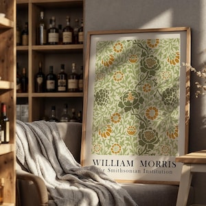 Op de afbeelding: Een ingelijste print met een bloemenpatroon in groen, oranje en crèmetinten. De print is voorzien van de tekst "WILLIAM MORRIS" en "the Smithsonian Institution". Het kunstwerk wordt getoond in een kamer met een houten boekenplank en een fauteuil.