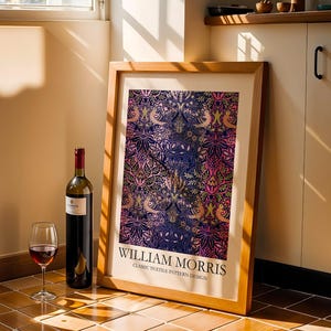 Op de afbeelding: Een ingelijste print met een klassiek textielpatroonontwerp van William Morris, met een rijk tapijt van kleuren, waaronder blauw, roze en goud. Een fles rode wijn en een wijnglas staan naast de lijst, op een betegelde vloer.
