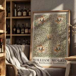Op de afbeelding: Een ingelijste print met een bloemmotief in groen, beige en oranje tinten. De print is voorzien van de tekst "WILLIAM MORRIS" en "The Smithsonian Institution". Het kunstwerk wordt getoond in een kamer met een houten plank en een stoel.