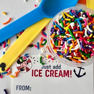 May include: A white card with a cartoon image of Mickey Mouse wearing a captain's hat. The card says "Just add ICE CREAM!" with a blue anchor symbol. The card is surrounded by colorful sprinkles and two plastic spoons, one blue and one yellow.