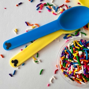 May include: Two plastic spoons with Mickey Mouse ears on the handles. One spoon is blue and the other is yellow. A small clear container filled with colorful sprinkles.