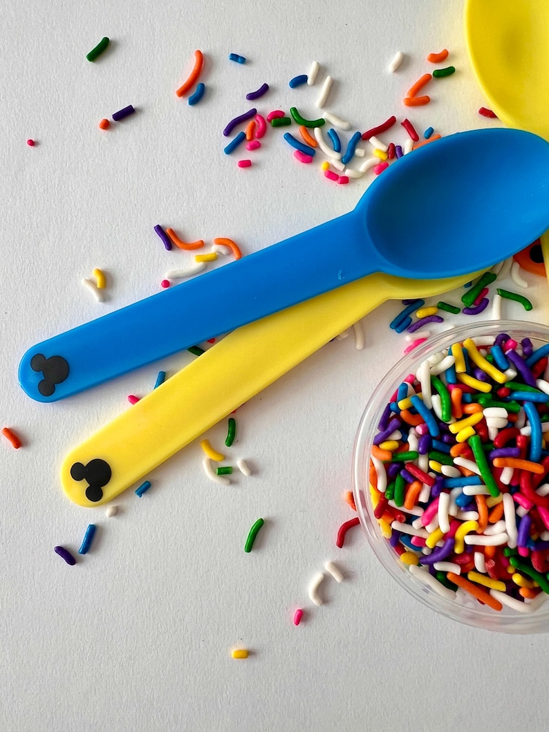 May include: Two plastic spoons with Mickey Mouse ears on the handles. One spoon is blue and the other is yellow. A small clear container filled with colorful sprinkles.