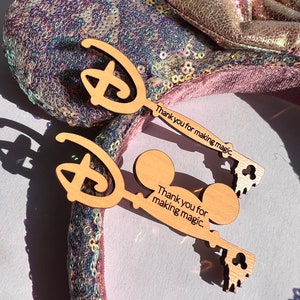 Könnte beinhalten: Zwei Schlüsselanhänger aus Holz in Form des Disney-Schlosses und von Mickey-Maus-Ohren. Die Schlüsselanhänger tragen den Text "Thank you for making magic."