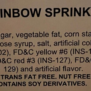 May include: A close-up of a white label with the words "Rainbow Sprinkles" printed in black. The label lists the ingredients, including sugar, vegetable fat, corn starch, soy lecithin, glucose syrup, salt, artificial colorants, and artificial flavor. It also states that the product is gluten-free, trans fat free, nut free, and contains soy derivatives.