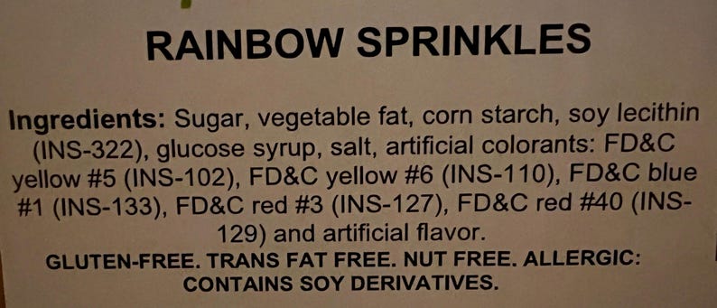 May include: A close-up of a white label with the words "Rainbow Sprinkles" printed in black. The label lists the ingredients, including sugar, vegetable fat, corn starch, soy lecithin, glucose syrup, salt, artificial colorants, and artificial flavor. It also states that the product is gluten-free, trans fat free, nut free, and contains soy derivatives.