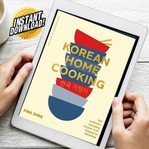 May include: A tablet displays a cookbook cover titled "KOREAN HOME COOKING" with stacked bowls in red, blue, and gold. The cover includes the text "INSTANT DOWNLOAD!" and "100 authentic everyday recipes, from bulgogi to bibimbap."