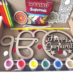 May include: A craft kit in a cardboard box, containing pencils, paint pots, and wooden cutouts. The kit includes a bag of Haribo Supermix sweets, stickers, and a colouring page. The wooden cutouts spell out "Happy Gurpurab".