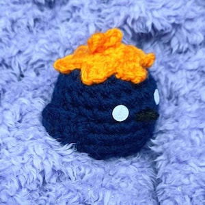 Könnte beinhalten: Ein handgehäkeltes blaues Vögelchen mit orangefarbenem Kamm, weißen Augen und einem schwarzen Schnabel. Der Vogel ist vor einem weichen, lavendelfarbenen, strukturierten Hintergrund platziert. Dieser skurrile Artikel ist perfekt für ein Kinderzimmer oder als Dekoration.