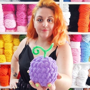 Könnte beinhalten: Eine gehäkelte lila Frucht mit einem grünen Stiel wird vor einem Regal voller bunter Wolle präsentiert. Die Frucht ist rund und strukturiert, mit einem grünen, lockigen Stiel. Der Hintergrund zeigt verschiedene Wollschattierungen, darunter Rosa, Gelb und Orange.