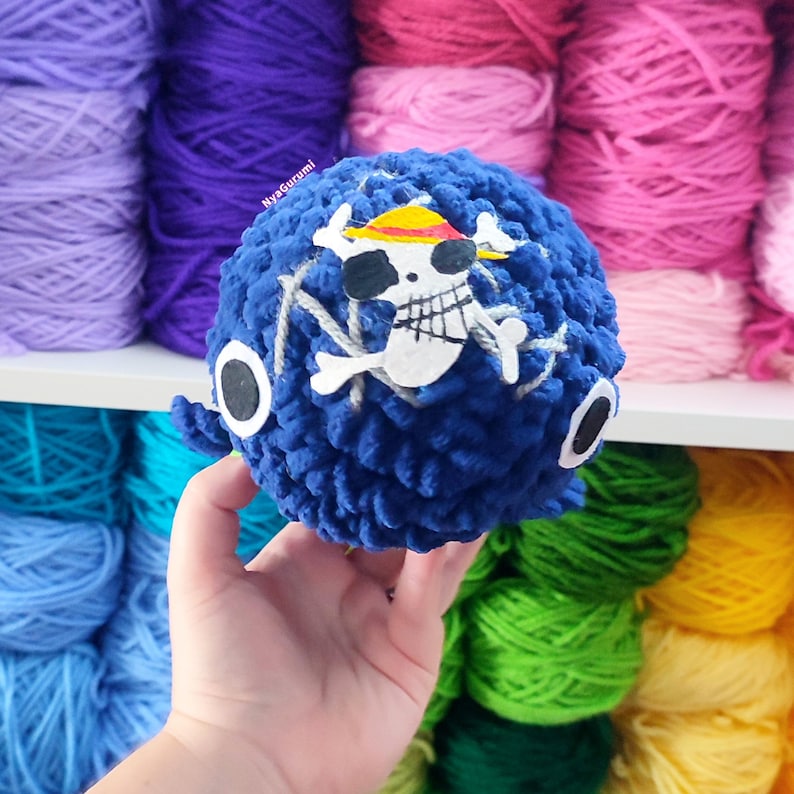May include: A handmade blue crochet ball featuring a white skull and crossbones design with a yellow and red hat. The ball has two black and white eye details. The background shows various colorful yarn skeins.