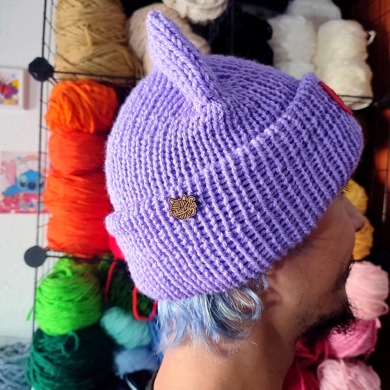 Op de afbeelding: Een paarse gebreide beanie met kattenoren en een klein houten pinnetje. De beanie is gemaakt met een textuur gebreide steek.