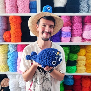 May include: A handmade blue crochet whale with white accents and a skull and crossbones design. The whale is held in front of a shelf filled with colorful yarn. A straw hat with a blue crochet creature is on top of the person's head.
