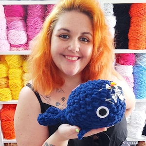 May include: A handmade, navy blue crochet whale with a white skull and crossbones design. The whale has a white eye with a black center. The background includes shelves filled with colorful yarn in shades of pink, yellow, white, and blue.