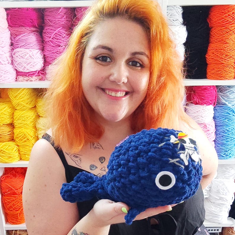 May include: A handmade, navy blue crochet whale with a white skull and crossbones design. The whale has a white eye with a black center. The background includes shelves filled with colorful yarn in shades of pink, yellow, white, and blue.