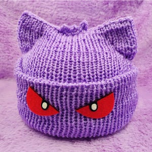 Op de afbeelding: Paarse gebreide beanie met rode ogen, die lijkt op een cartoonfiguur.