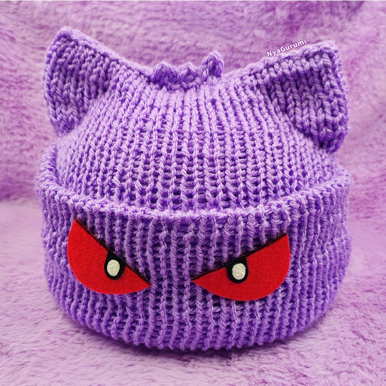 Op de afbeelding: Paarse gebreide beanie met rode ogen, die lijkt op een cartoonfiguur.