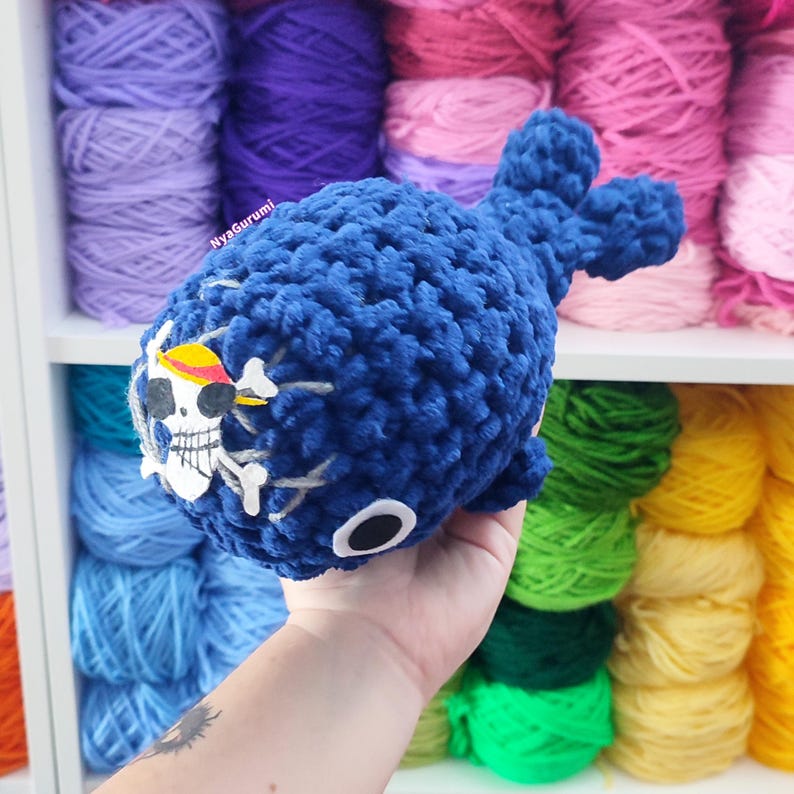 May include: A handmade blue crochet whale with a skull and crossbones design on its side. The whale has a round body, a tail, and two small fins. The skull is white with a yellow hat and the crossbones are white. The background shows colorful yarn.