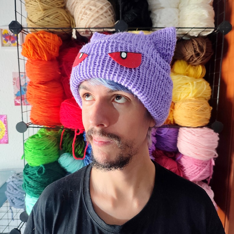 Op de afbeelding: Een persoon draagt ​​een paarse gebreide muts met rode ogen. De muts heeft een kattenoorontwerp. De persoon heeft blauw haar en een baard. De achtergrond is een muur met planken vol kleurrijke wol.