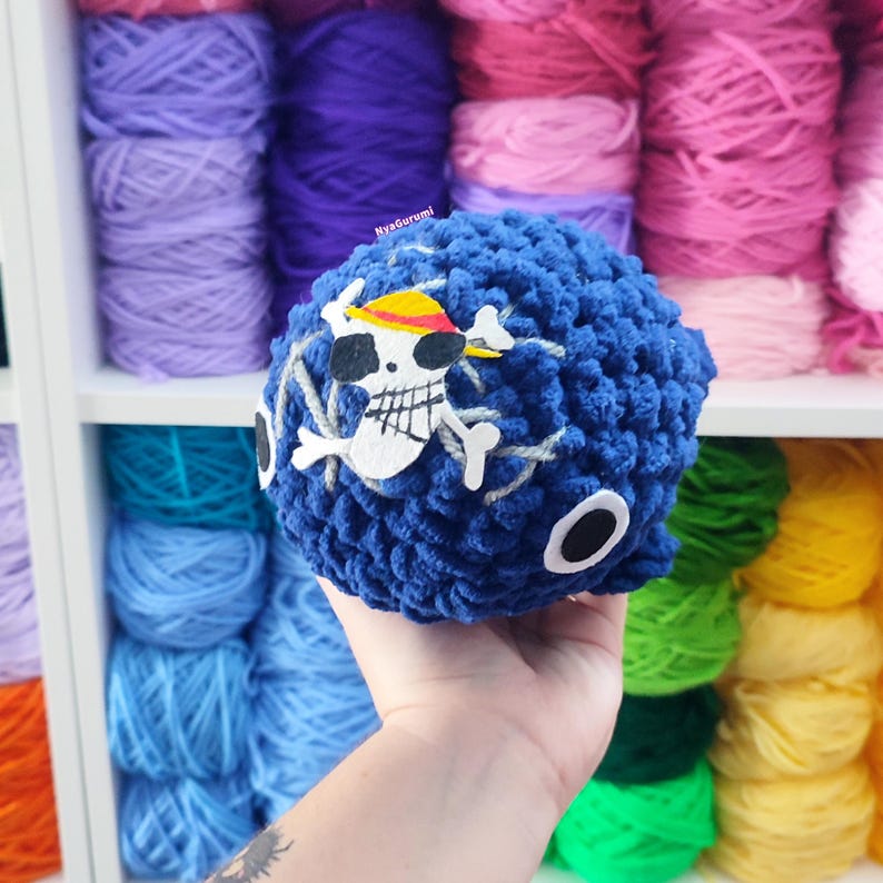 May include: A blue crocheted ball with a white skull and crossbones design, featuring a yellow and red hat. The ball has white accents and is held in front of a shelf filled with colorful yarn.