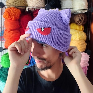 May include: A purple knitted beanie with cat ears and red felt eyes. The beanie is being worn by a person with a beard.