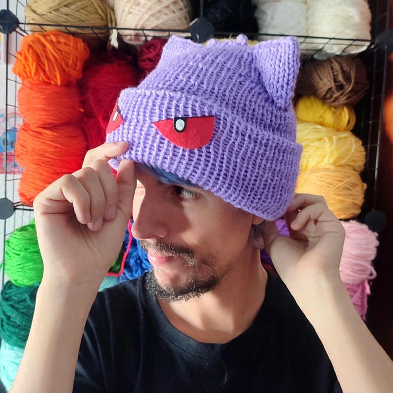 May include: A purple knitted beanie with cat ears and red felt eyes. The beanie is being worn by a person with a beard.