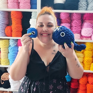 May include: A woman holds two handmade blue crochet toys, a small round one and a larger whale-shaped one. The whale has white accents and a large eye. The background features shelves filled with colorful yarn skeins, including orange, pink, and blue.