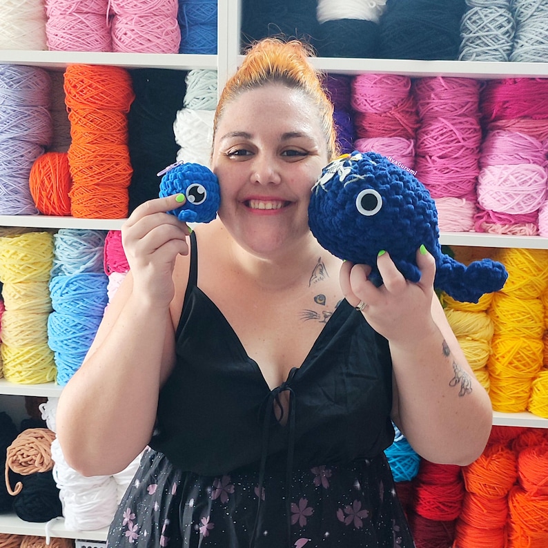 May include: A woman holds two handmade blue crochet toys, a small round one and a larger whale-shaped one. The whale has white accents and a large eye. The background features shelves filled with colorful yarn skeins, including orange, pink, and blue.