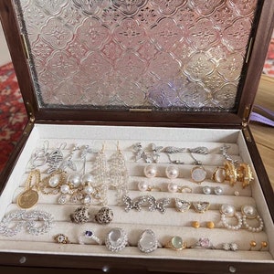 Puede incluir: Una caja de joyería de madera con una tapa de vidrio y un diseño estampado. La caja está abierta y llena de varios pendientes, anillos y collares. La joyería está hecha de oro, plata y perlas.