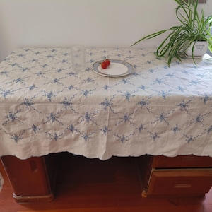 以下が含まれることがあります： 青い花柄の白いテーブルクロスが木製の机を覆っています。透明なグラス、赤いトマトが2つ乗ったお皿、鉢植えの植物がテーブルにあります。テーブルクロスはスカラップの縁取りです。
