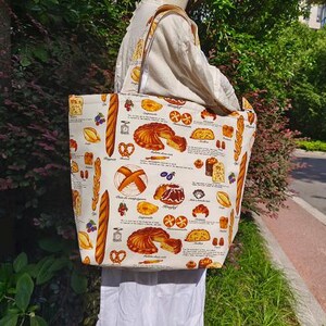 Puede incluir: Una bolsa de lona blanca con un diseño colorido de varios panes y pasteles. La bolsa tiene un asa de cuero marrón y la lleva una persona.