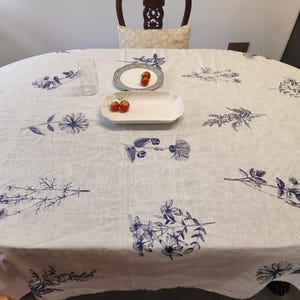 Puede incluir: Un mantel ovalado blanco con un estampado floral azul. El mantel está sobre una mesa de madera con un vaso y un plato con tomates. El mantel tiene un estilo vintage y rústico.