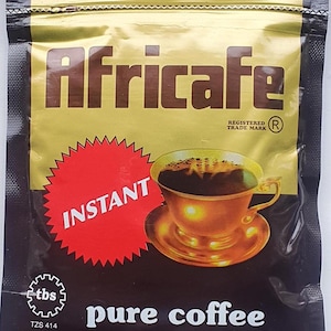 以下が含まれることがあります： アフリカカフェのインスタントピュアコーヒーの密封されたパッケージ。パッケージは主に黒と金で、ブランド名が大きな茶色の文字で書かれています。「INSTANT」と書かれた赤いバーストが前面にあり、コーヒーカップのイラストも描かれています。