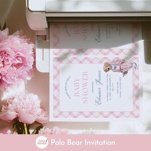 May include: A baby shower invitation featuring a pink plaid border and a watercolor illustration of a teddy bear wearing a pink outfit. The invitation includes the text "Baby Shower" and event details. Pink peonies are in the background.