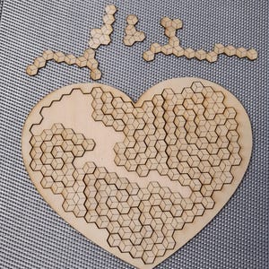 Può includere: Un puzzle di legno a forma di cuore con un design geometrico. Il puzzle è composto da piccoli pezzi che si incastrano tra loro per formare un motivo di cubi. Al puzzle mancano alcuni pezzi, che sono mostrati separatamente su una superficie grigia.