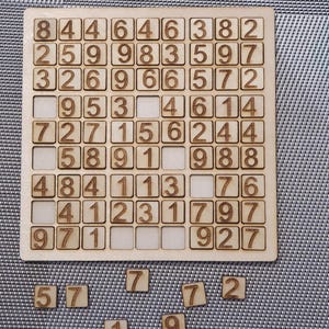 Può includere: Un tabellone di gioco in legno con una griglia di 9x9 quadrati, ogni quadrato contiene un numero. I numeri 5, 3, 7, 2, 1 e 9 sono anche visibili sul tabellone.