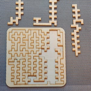 Może przedstawiać: Drewniana układanka z kwadratową ramą i kilkoma mniejszymi elementami. Elementy są ułożone w siatkę i mają jasnobrązowy kolor.