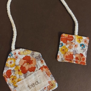 May include: Floral tea bag tags with a white cord. The tags are rectangular and feature a colorful pattern of orange, yellow, and blue flowers on a white background. The word "tea" is printed on a white label.