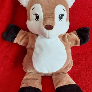 Puede incluir: Una marioneta de mano de peluche con forma de ciervo, con pelaje marrón, vientre blanco y marcas blancas en la cara. La marioneta presenta pezuñas negras en los brazos y piernas, orejas blancas esponjosas y ojos bordados con pestañas largas.