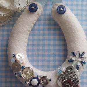 Puede incluir: Un herradura de fieltro blanco decorada con botones azules, cuentas y un dije de corazón plateado. El dije lleva la inscripción "You are always in my heart". Se adjunta una cinta azul claro para colgar.
