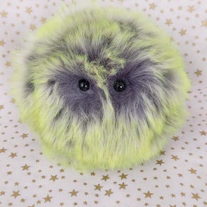 May include: A round, plush, fuzzy creature with long, soft faux fur in vibrant neon yellow and dark grey. It features two small, round, black, glossy eyes embedded within the dense, shaggy texture of the spherical, stuffed toy.