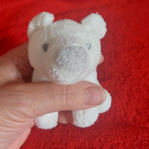 May include: A small, white plush polar bear toy with a soft, textured fabric surface. The bear features two small, dark grey embroidered eyes and a rounded, slightly darker grey nose. The ears are small and rounded, positioned on top of the head.